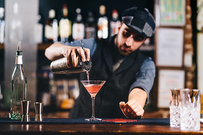 Male bartender with fedora pours cocktail into glass